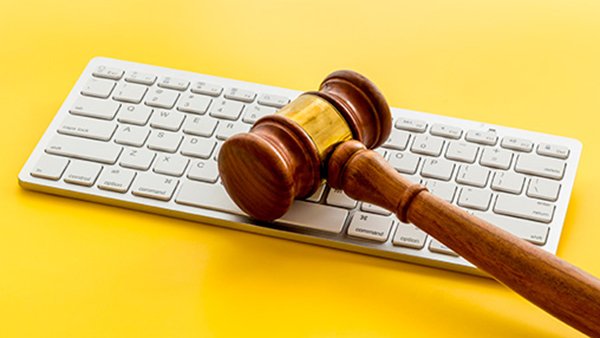 Tietokoneen näppäimistö, jonka päällä lepää puisesta ja metallisesta osasta koostuva puheenjohtajan nuija. Tausta on kirkkaan keltainen.